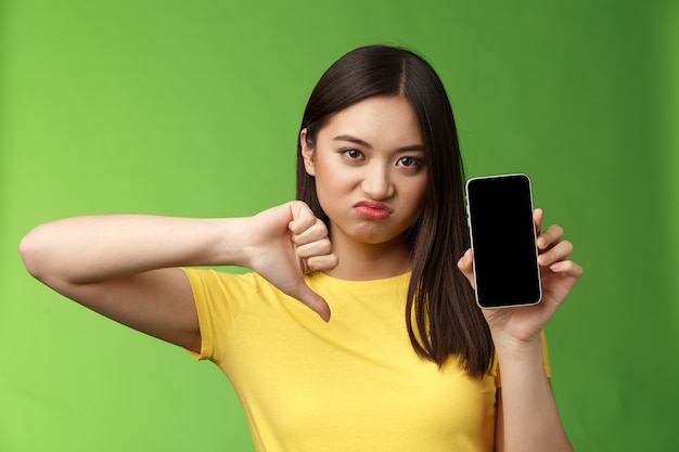 クローズアップ失望した動揺したアジアの女の子が悪いひどいアプリを判断し、スマートフォンの画面を表示し、親指を下に向けて顔をゆがめ、否定的なフィードバックを与え、元ボーイフレンドの新しいガールフレンドに耐えられない