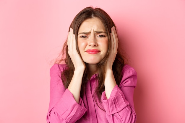 Close up of distressed and bothered young woman covering her ears, grimacing cant bear this anymore, standing overwhelmed against pink wall.