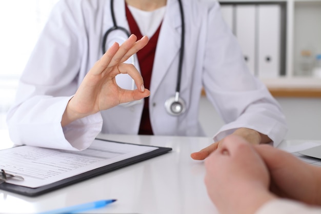 Close up of a doctor hand showing ok sign while phusician and his patient discussing medical records after health examination.