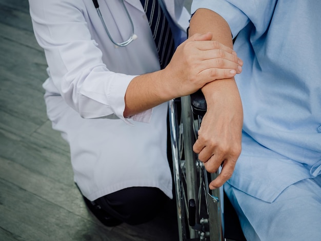 Primo piano le mani del medico che tengono la mano del paziente anziano su una sedia a rotelle il medico maschio in abito bianco visita e incoraggia il paziente anziano vestito di azzurro in ospedale concetto di assistenza agli anziani