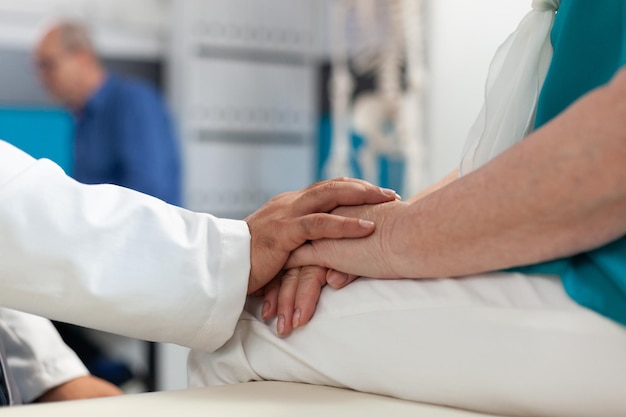 Крупным планом - доктор, поддерживающий пенсионерку с физической болью, держась за руки пациента, чтобы обеспечить выздоровление и лечение. Медик утешает пожилого человека с артритом на физиотерапии