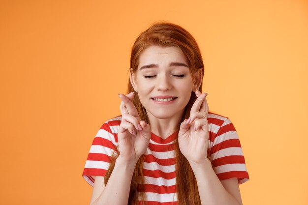 Close-up dreamy cute tender ginger girl make wish, cross fingers good luck closed eyes bite lip eager win lottery, supplicating, praying dream come true, stand orange background believe miracle