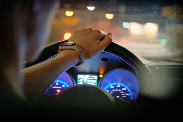 Close up of driver hands holding steering wheel driving car with blurred city street lights on background at night