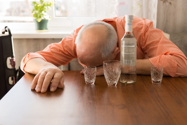 Close-up dronken kale ouderen die een dutje doen op de houten tafel met wodka en kleine glazen.