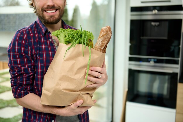 Close-up eco paper bag, craft cardboard packet with greens,\
salads, healthy food, whole grain baguette bread in hands of\
handsome caucasian guy smiling beautiful toothy smile standing in\
home kitchen