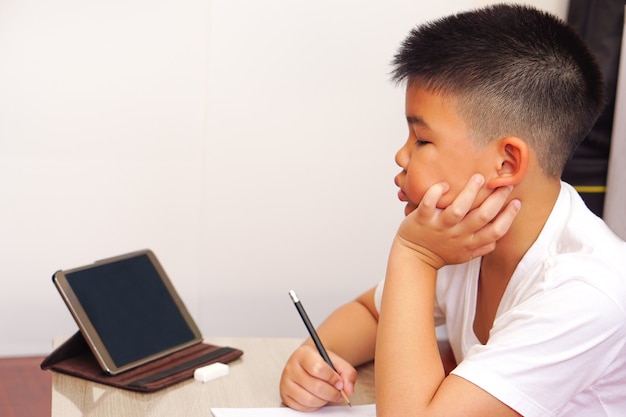 Close-up een aziatische jongen in een wit t-shirt die naar digitale tablet kijkt, zoekt de informatie (kind denkt) en doet huiswerk of schrijft notitieboekje met potlood op tafel.