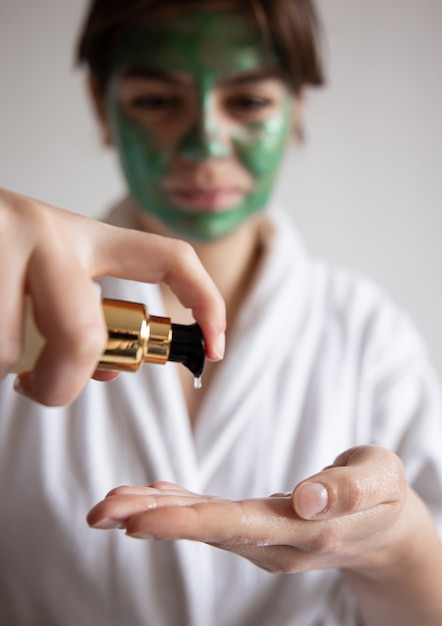 Close-up, een vrouw in een wit gewaad met een groen cosmetisch masker drukt op de dispenser van schoonheidsverzorgingsproducten, onscherpe achtergrond.