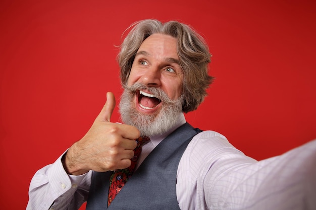 Close up of elderly gray-haired mustache bearded man in shirt vest and tie isolated on red background. People lifestyle concept. Mock up copy space. Doing selfie shot on mobile phone showing thumb up.