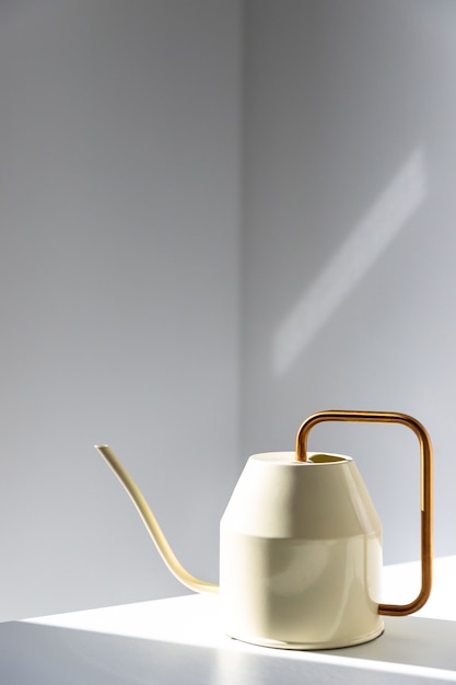 Close up of elegant white metallic watering can with long thin spout and yellow thin circular handle on the table, wall on surface, indoors. Sunlight.