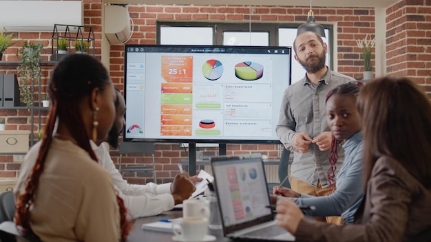 Close up of employee showing business development on charts presentation at briefing meeting with people. Man presenting company growth and data analysis on monitor in boardroom.