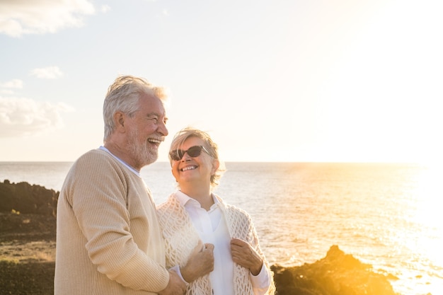 Close-up en portret van twee gelukkige en actieve senioren of gepensioneerden die plezier hebben en genieten van het kijken naar de zonsondergang die lacht met de zee - oude mensen die buiten samen van vakanties genieten