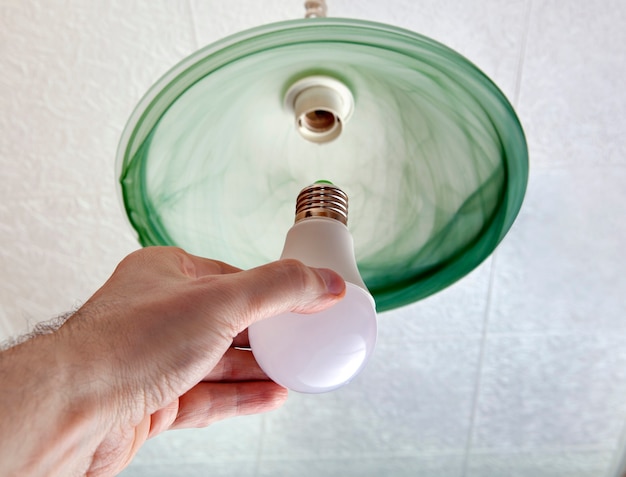 Close-up of energy-saving LED light bulb in human hand, replacement of  lamp in ceiling luminaire made of green ground glass.