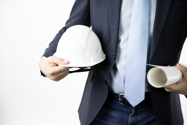 Close up engineer in formal suit holds hard safety helmet hat and blueprint roll