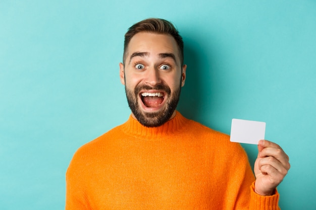 Close-up of excited caucasian man showing his credit card, smiling and staring amazed, standing in orange sweater against turquoise background.