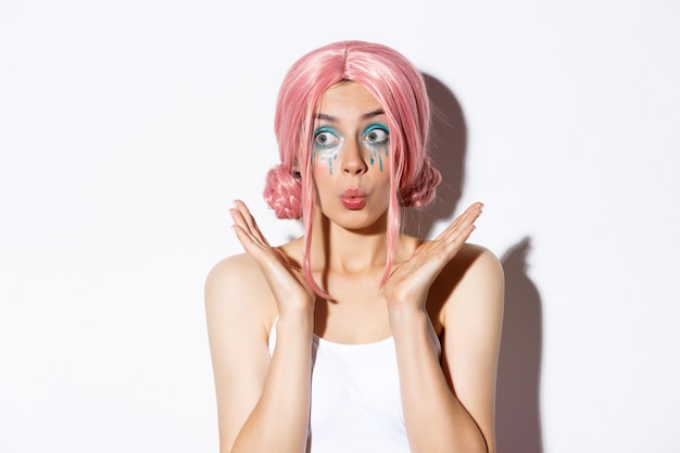 Close-up of excited party girl in pink wig and bright makeup looking impressed, smiling and gazing surprised, standing over white background