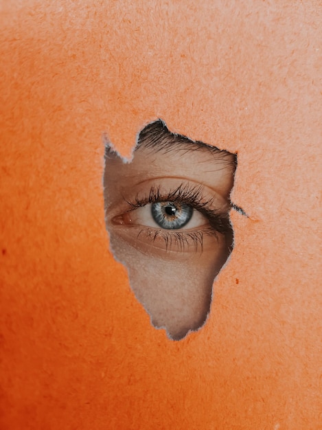 Photo close-up eye peeking through torn orange paper