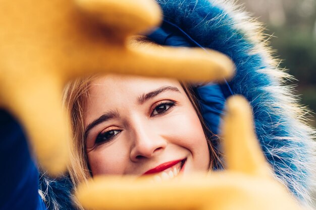 Close-up of the face of a beautiful young adult woman in\
winter, wearing a blue coat, yellow gloves and red lipstick, making\
the symbol of a photo frame with her hands. looking to the\
camera.