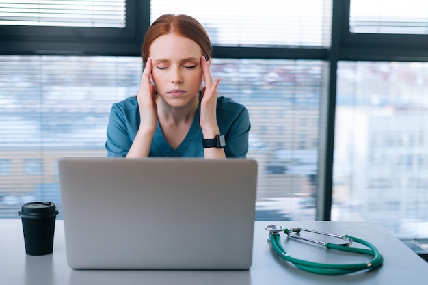 Close-up face of exhausted tired young female doctor in blue green medical uniform massaging temples, suffering from headache sitting on background of window in office.