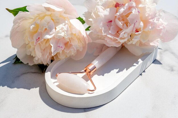 Primo piano del rullo per massaggio facciale su vassoio in pietra e sfondo in marmo bianco concetto di cura di sé e di bellezza decorazione di fiori di peonia rosa