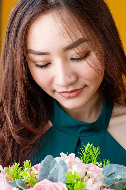 Close up face shot of cute and pretty curly hair Asian female brunette holding flower bouquet with cheerful and happy, studio shot isolated on bright yellow background.