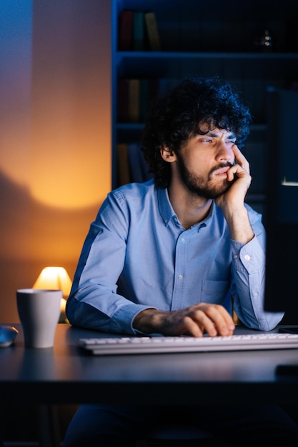 Close-up face of sleepy young bearded man working late at night on computer rubbing eyes at home office. Tired sleepy student prepares for late night session. Freelancer working late over project.