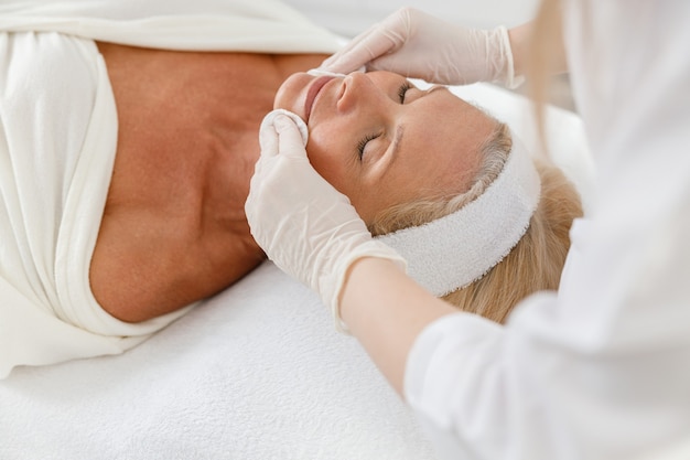 Foto primo piano viso spa trattamento di bellezza, cura della pelle. donna maggiore che ottiene cura del viso dall'estetista al salone della stazione termale, vista laterale.