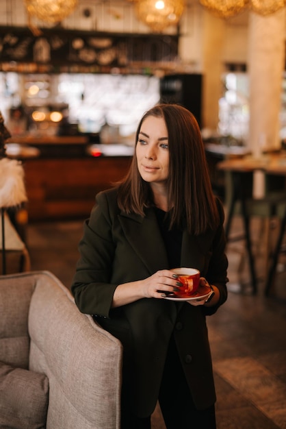 Крупный план лица молодой женщины, держащей в руке чашку вкусного горячего кофе, стоя в ресторане Кавказская дама позирует со стаканом напитка в кафе с современным темным интерьером