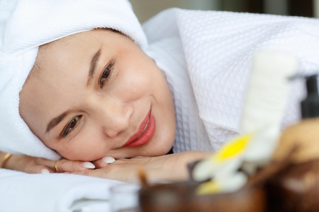 Close up facial shot of asian happy female model in white clean\
bathrobe lying down closed eyes relaxing on massage bed with\
compress ball coconut oil and plumeria in tray smiling wait for\
therapy.