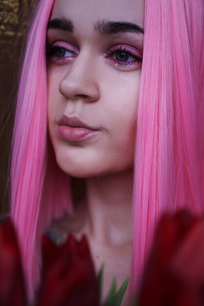 Close up fashion portrait. Vertical photo. Portrait of a dreamy girl with bright pink hair with flowers close to face. Looking to the side