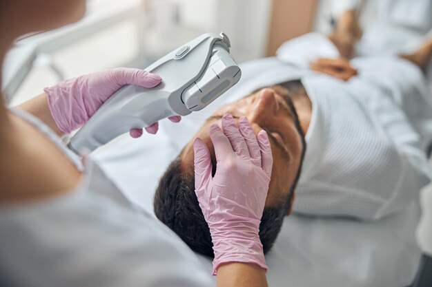 Close up of female beautician hands in sterile gloves holding laser device and touching male forehead