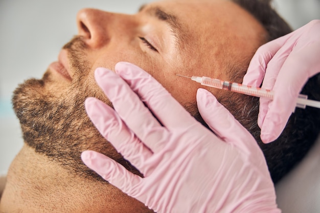 Close up of female beautician hands in sterile gloves making injection into male face