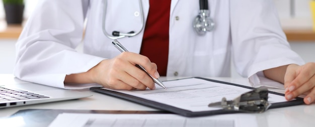 Close up of a female doctor filling up  an application form while sitting at the table. Medicine and health care concept