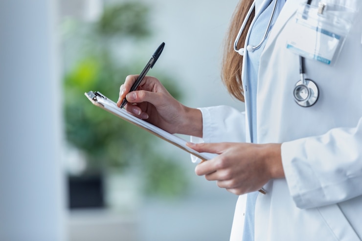 Photo close-up of a female doctor filling up medical form at clipboard while standing in the consultation.