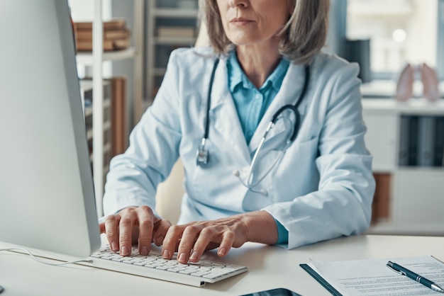 Close up of female doctor in white lab coat working using computer while sitting in her office