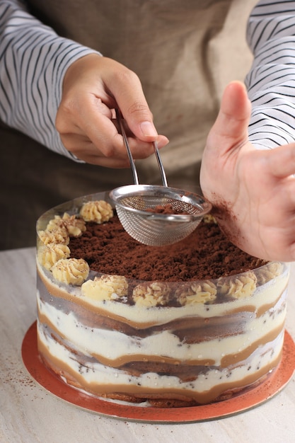 Close Up Female Hand Dusting Tiramisu like Cake with Cocoa Powder. Still life with Whole of Cake  on Wooden Table.