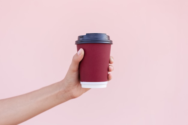 Close-up of female hand holding a paper coffee cup on the background of pastel pink color.