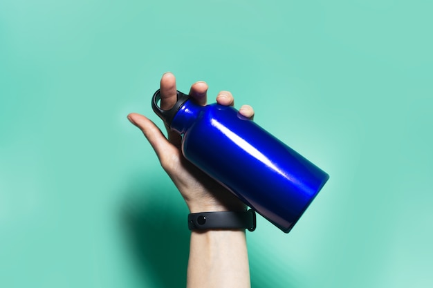 Close-up of female hand holding a reusable eco, metal thermo water bottle of phantom blue color, isolated on aqua menthe