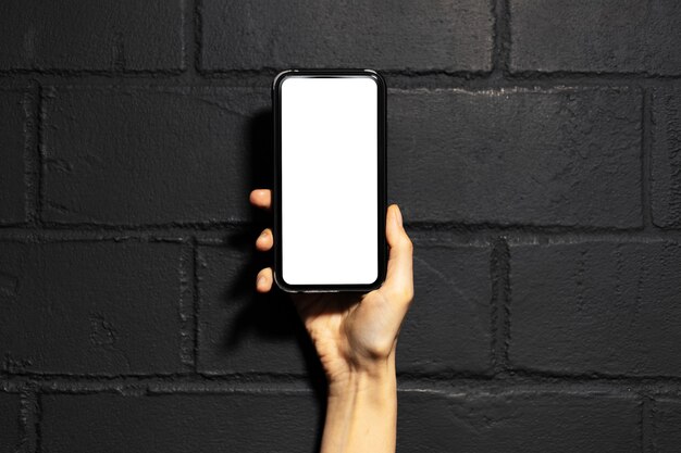 Close-up of female hand holding a smartphone with mockup, on the background of black brick wall.