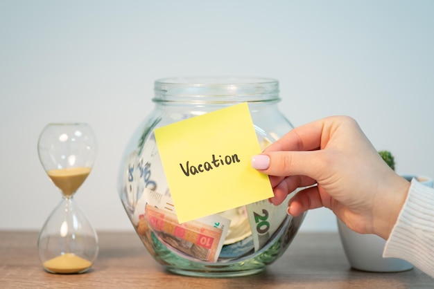 Close up of female hand holding sticker with word Vacation on it glass moneybox and sandglass on the background Financial planning saving for pleasure and relax on a vacation concept