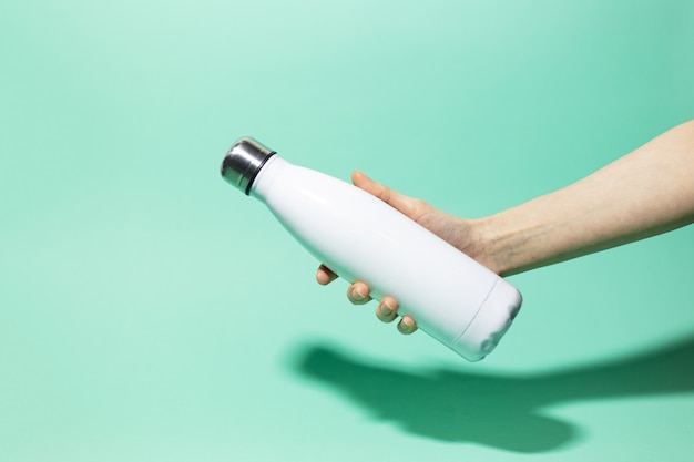 Крупный план женской руки, держащей белую многоразовую стальную нержавеющую термо-бутылку с водой, изолированную на стене цвета морской волны. Без пластика.
