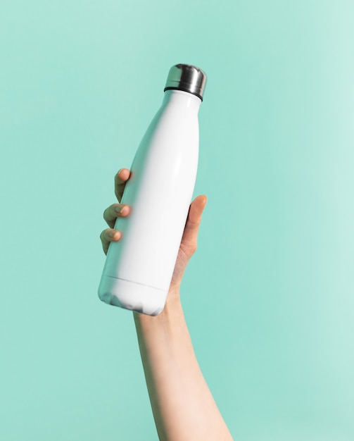 シアン、アクアメンテ色の壁に分離された白い再利用可能な鋼ステンレスサーモウォーターボトルを持っている女性の手のクローズアップ。プラスチックフリー。