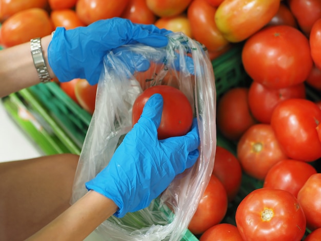 Close-up. Female hands in blue gloves choosing a tomato in supermarket during pandemic coronavirus COVID-19.