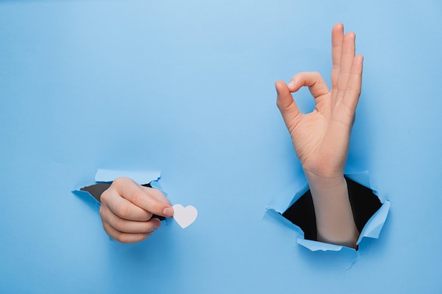 Close up of female hands holding little white hearts through and shows an ok sign a torn blue paper wall.