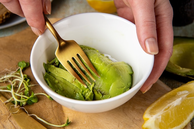Close up of female hands mashing avocado with fork in white\
plate
