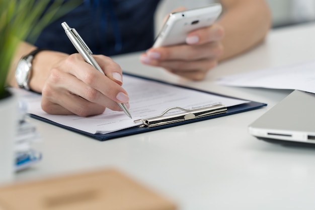 Primo piano delle mani femminili. donna che scrive qualcosa e guardando lo schermo del telefono cellulare seduto nel suo ufficio