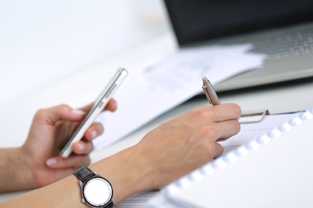 Primo piano delle mani femminili che lavorano con i documenti in ufficio. donna che scrive qualcosa e che guarda lo schermo del telefono cellulare