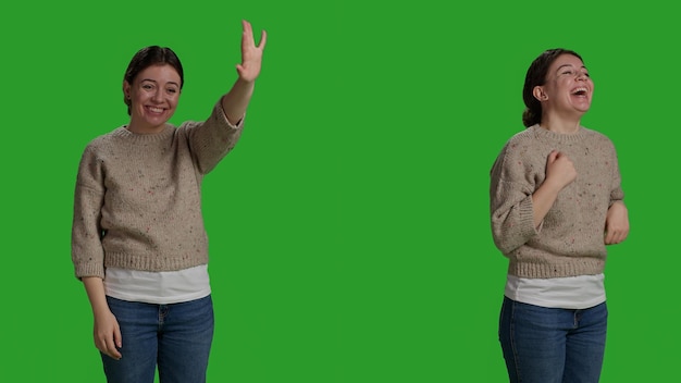 Close up of female model greeting someone and waving, smiling in studio. Relaxed young woman waving hi or hello on camera, standing over full body greenscreen backdrop and saying goodbye.