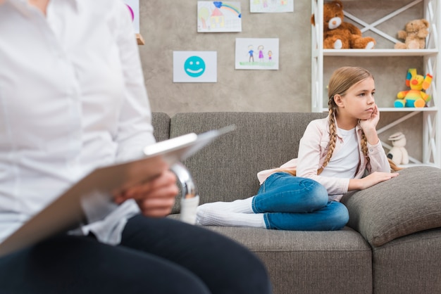 Close-up of female psychologist holding clipboard sitting in front of depressed girl