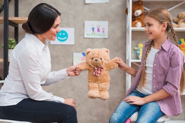 Close-up of female psychologist and smiling girl both holding teddybear in hands