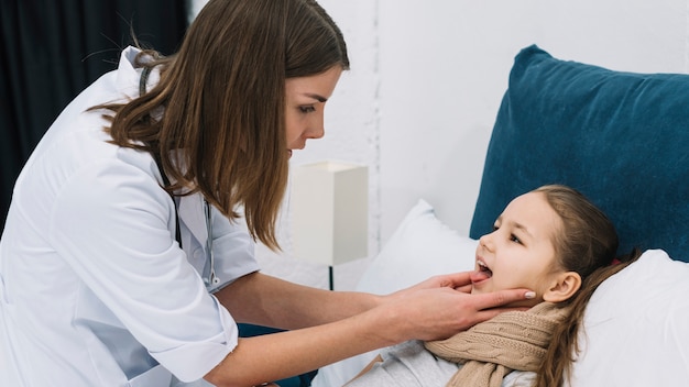 Foto primo piano del medico della femmina che controlla la gola di una ragazza ammalata che si trova sul letto